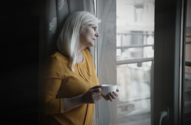 Starsza smutna kobieta stojąca przy oknie z filiżanką w ręce